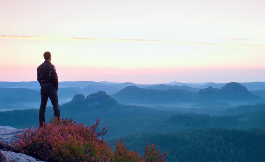 A man standing on top of a mountain. He is over looking a valley with mountains and a pink sunset.